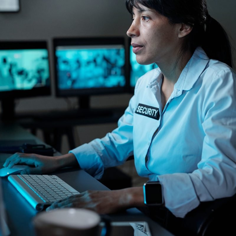 Young serious woman in security uniform concentrating on watching cctv on computer screen while sitting by workplace in control room