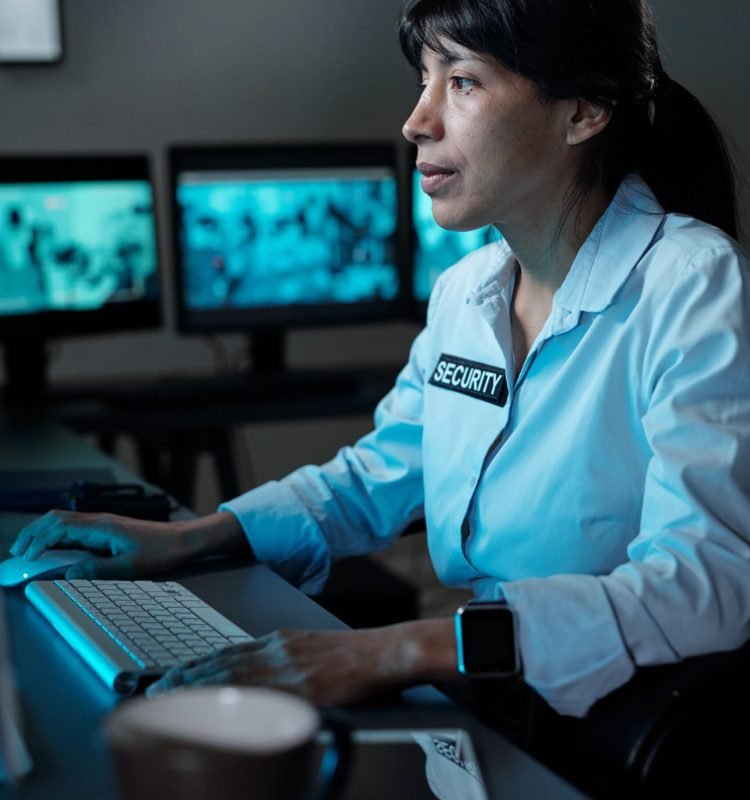 Young serious woman in security uniform concentrating on watching cctv on computer screen while sitting by workplace in control room